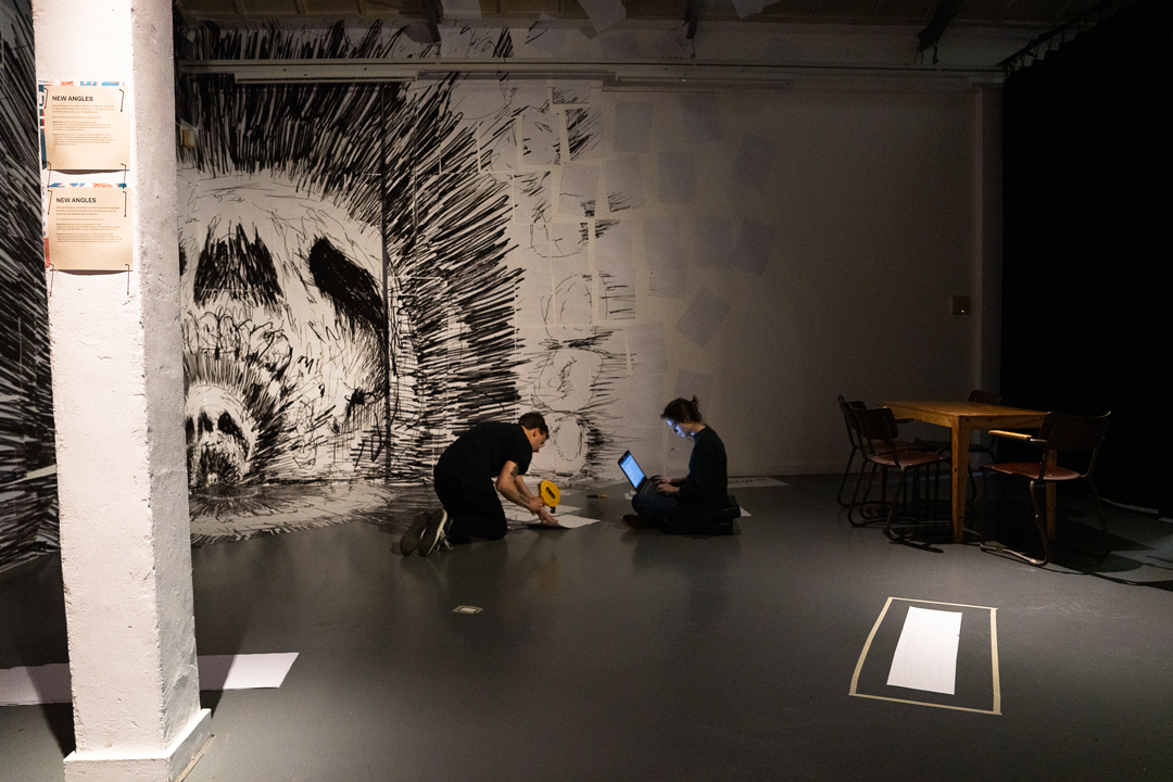 Kunstenaar wordt geïnterviewd terwijl hij aan het werk is in een kunstgalerie. We zien hem een grote tekening in delen aan een muur plakken.
